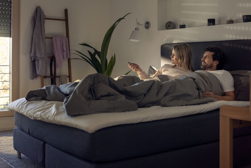 Paar liegt im Bett und öffnet per Fernbedienung die Rollläden am Morgen