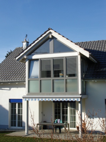 Weißes Haus mit Giebelgaube, Balkon und gestreifter Markise unter blauem Himmel.