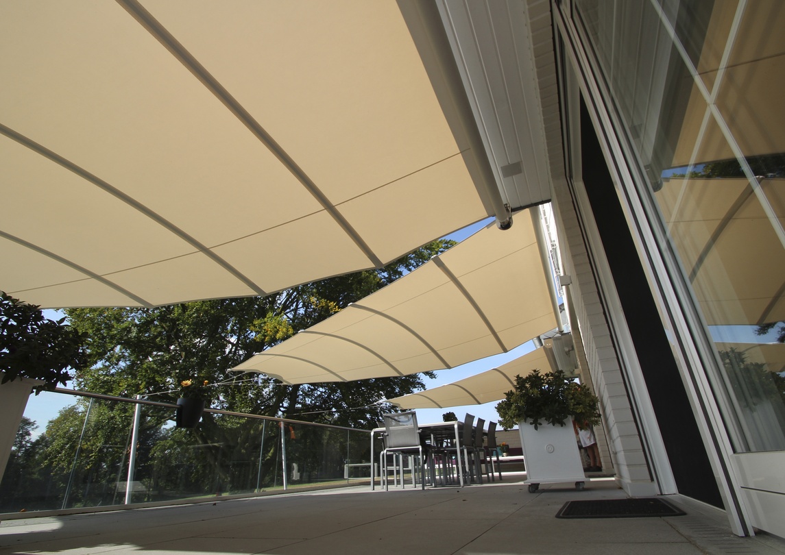 Helle Terrasse mit mehreren beigen Sonnensegeln, Sitzbereich und Gebäudefassade.