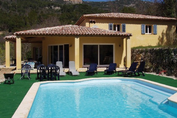 Gelbes Haus mit Pool, Liegestühlen und Grill vor bewaldeten Bergen.