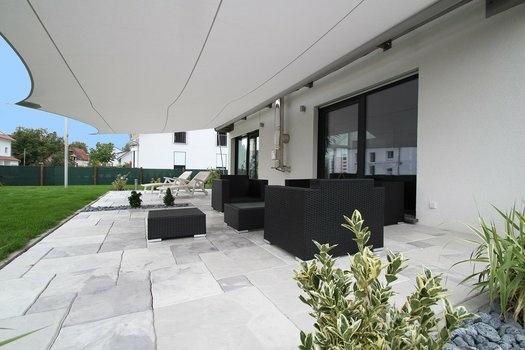 Moderne Terrasse mit hellen Steinplatten, schwarzem Mobiliar und weißem Sonnensegel.