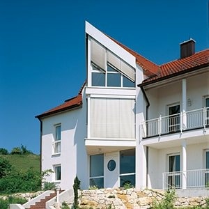 Modernes weißes Haus mit dreieckiger Glasfassade, Außenjalousien und rotem Dach.
