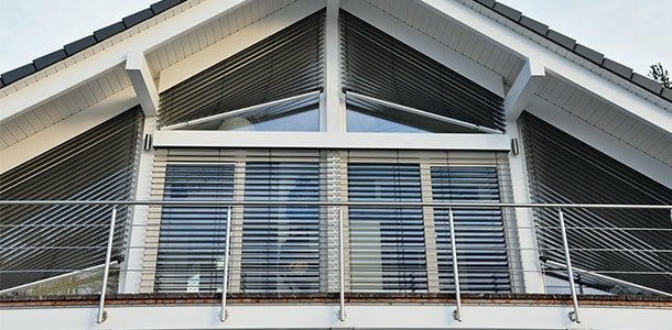 Moderner Hausgiebel mit Fenstern, teilweise geschlossenen Raffstores und Balkon mit Metallgeländer.