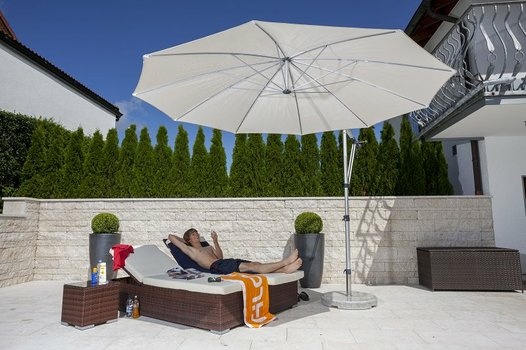 Mann entspannt auf Sonnenliege unter großem weißen Sonnenschirm auf sonniger Terrasse.