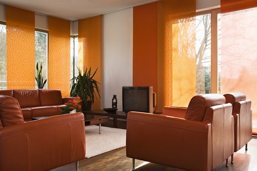 Modernes Wohnzimmer mit orangefarbenen Schiebevorhängen, braunem Ledersofa und Sesseln. Pflanzen und TV.