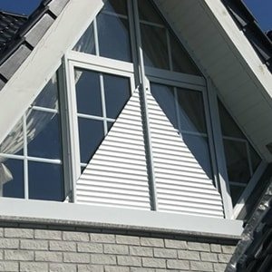Weißes, dreieckiges Giebelfenster mit Lamellen über heller Ziegelmauer und dunklem Dach.