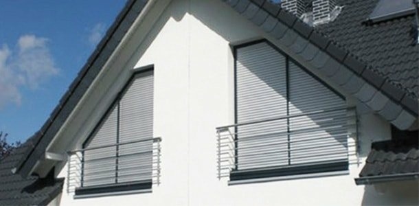 Dachgiebel eines modernen Hauses mit zwei dreieckigen Fenstern, hellgrauen Rollläden und Stahlgeländern.