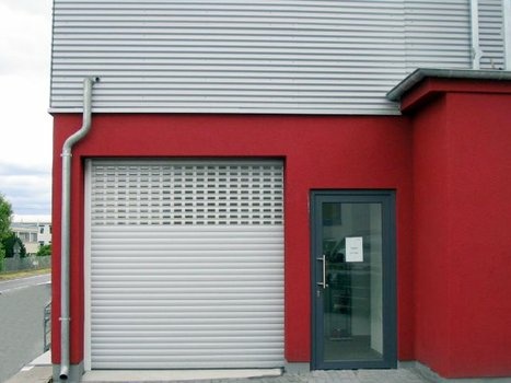 Rotes Gebäude mit grauem Wellblech, Rolltor und gläserner Eingangstür.