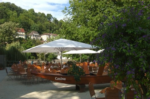 Gartenrestaurant mit weißen Sonnenschirmen, besetzten Tischen und einem Holzboot mit „Casino“.