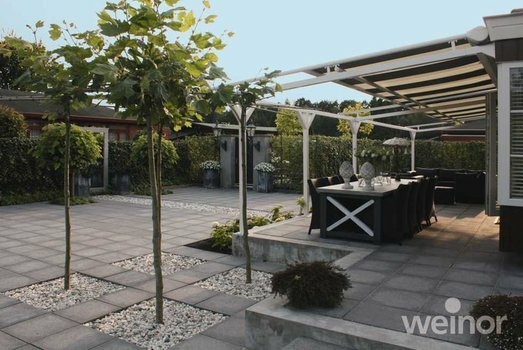 Große moderne Terrasse mit grauen Steinplatten, Bäumen und Essbereich unter weißer Pergola.