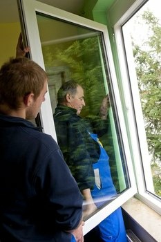Zwei Männer in blauen Overalls montieren einen neuen weißen Fensterrahmen.