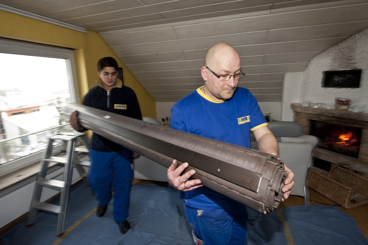 Zwei Arbeiter tragen eine schwere Rollladenkomponente durch einen Raum mit Kamin.
