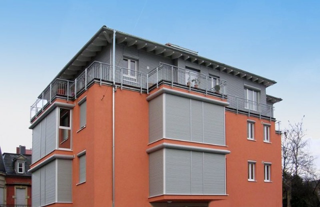 Fotografie: Modernes Wohnhaus mit orangeroter Fassade, grauen Rollläden und Balkonen.