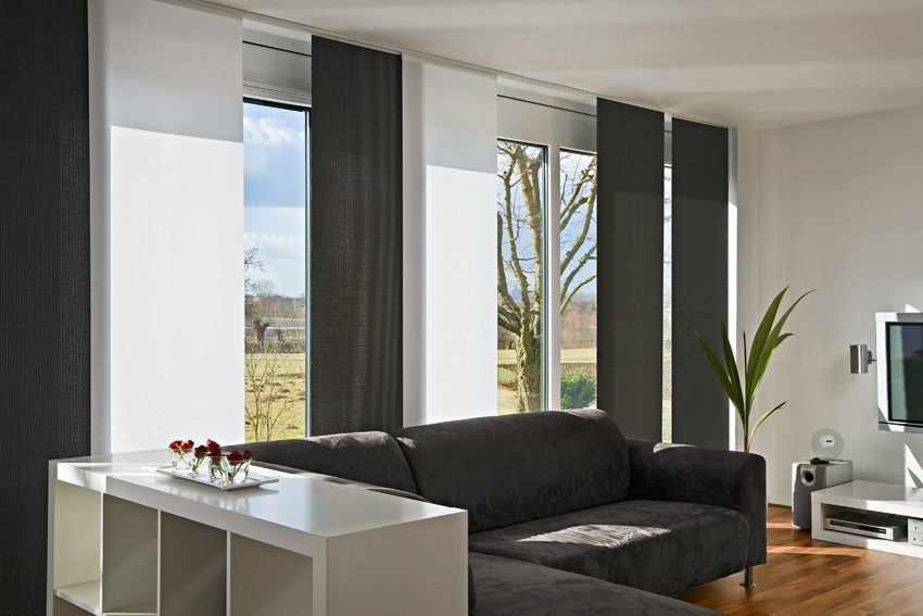 Modernes Wohnzimmer mit weißen und dunkelgrauen Flächenvorhängen, dunklem Sofa und Blick ins Grüne.