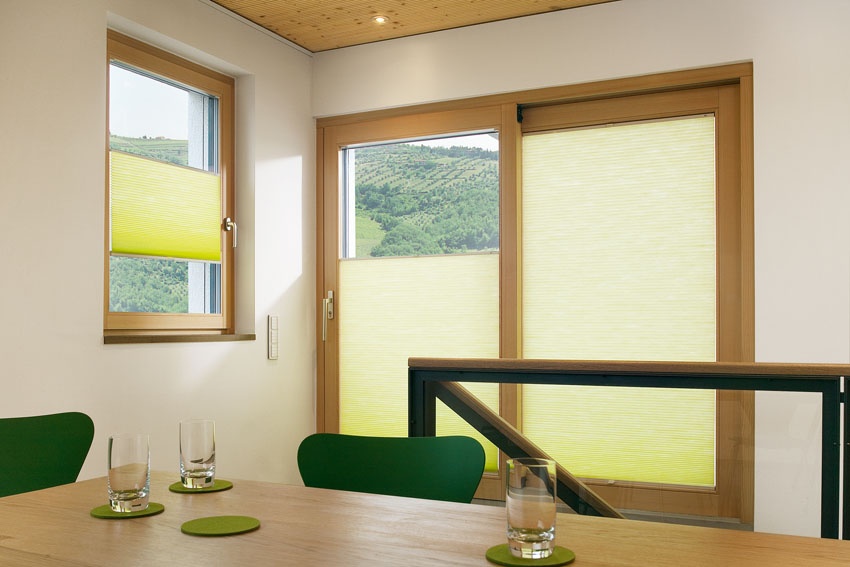 Helles Zimmer mit Holzschiebetür und Fenster, gelb-grünen Plissees, Holztisch, Bergblick.