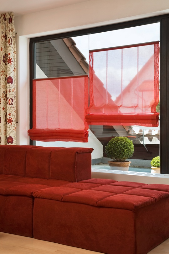 Modernes rotes Sofa an Fensterfront mit roten Plissees, draußen grüne Pflanze.