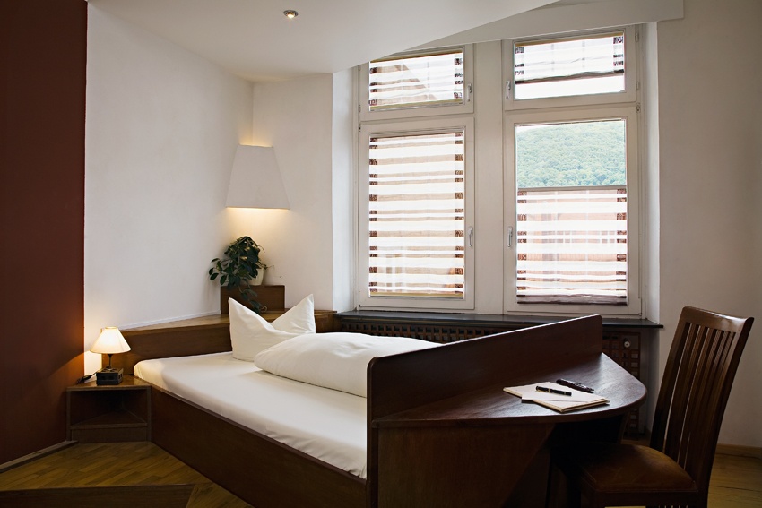 Gemütliches Einzelzimmer mit Bett, Schreibtisch und Fenster mit Jalousien.