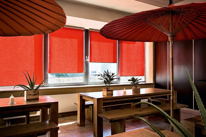 Gemütliches Restaurant mit roten Rollos an großen Fenstern, Holztischen und Deko-Sonnenschirmen.