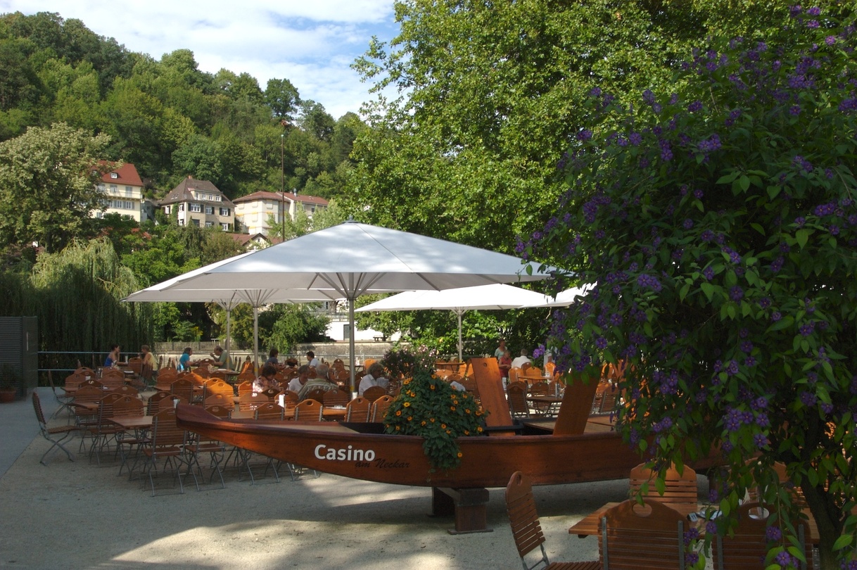 Belebter Biergarten mit Sonnenschirmen und einem Holzboot. Grüne Bäume und Häuser im Hintergrund.