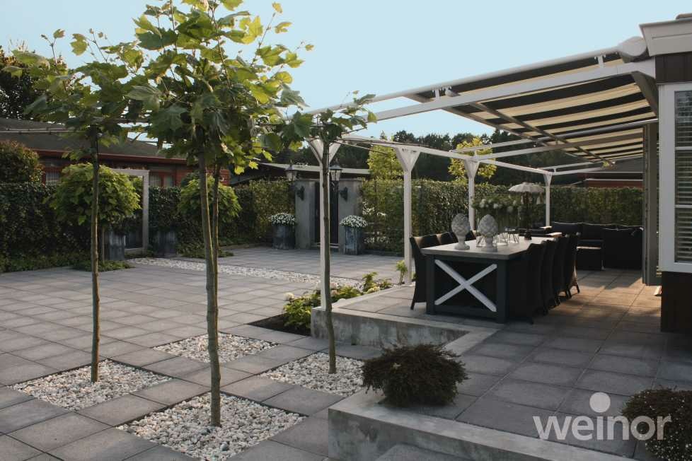 Moderne Terrasse mit grauen Pflastersteinen, kleinen Bäumen und überdachtem Essbereich unter weißer Pergola.