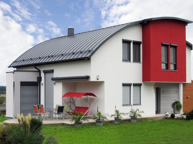 Modernes Haus mit geschwungenem Dach, weißen Wänden und rotem Akzent. Grüner Garten.