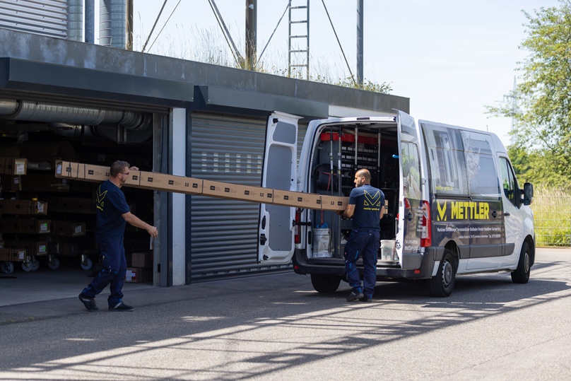 Zwei Männer verladen eine lange Leiter in einen weißen Transporter bei Sonnenschein.