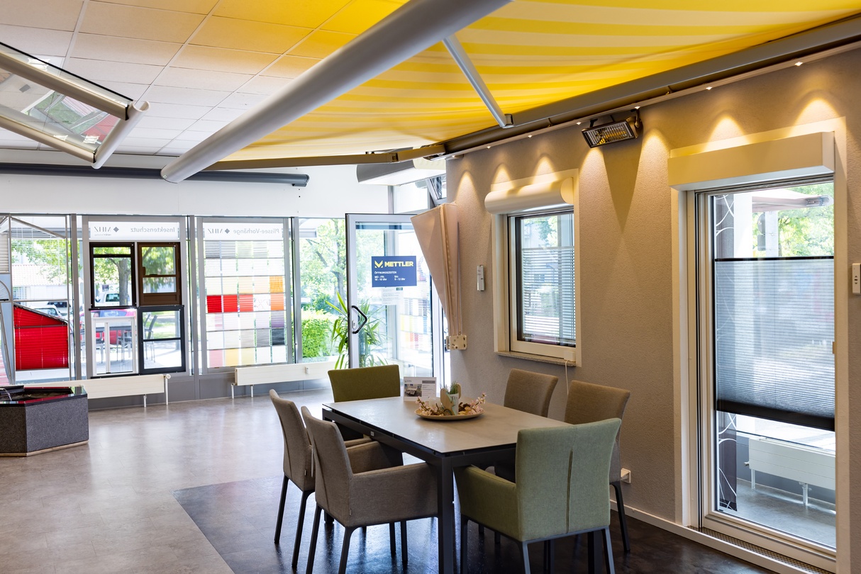 Showroom mit Fenstern, gelber Markise und Esstisch mit Stühlen.