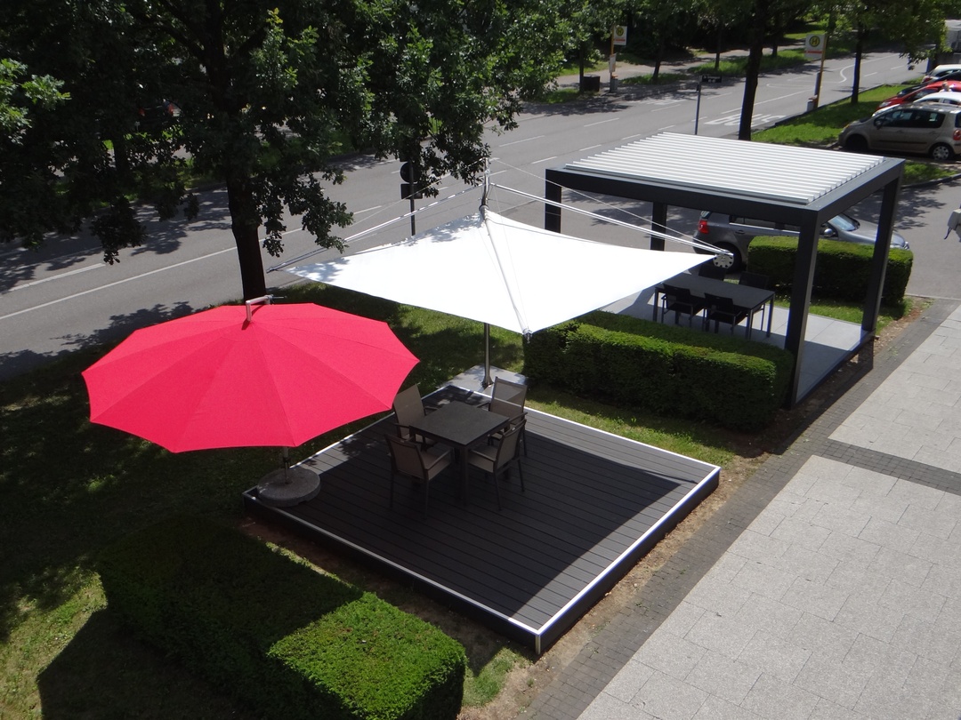 Sonnige Außenterrasse mit rotem Schirm, weißem Sonnensegel und Pergola über Sitzgelegenheiten.