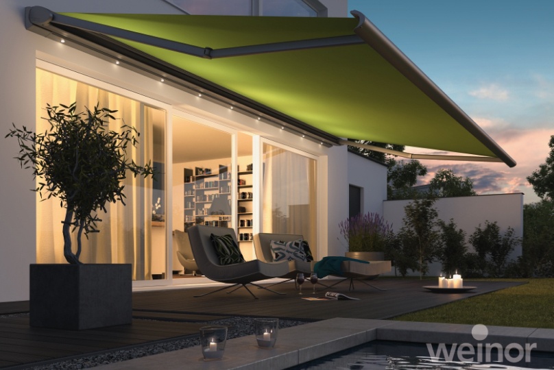 Moderne Terrasse bei Dämmerung mit großer grüner Markise, warmer Beleuchtung, Gartenmöbeln und Pool.