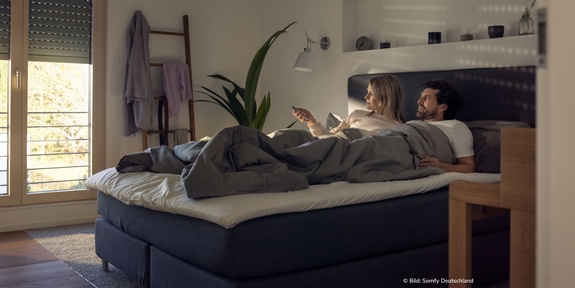 Paar liegt im Bett und öffnet per Fernbedienung die Rollläden am Morgen