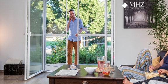 Mann telefoniert am offenen Balkon, Person entspannt im Wohnzimmer.