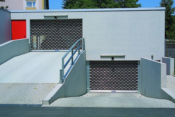 Eingang einer Tiefgarage mit zwei Rollgittertoren, einer Rampe und roter Tür.