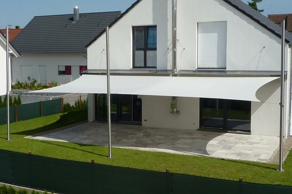 Modernes weißes Haus mit großem Sonnensegel über gepflasterter Terrasse und Rasen.