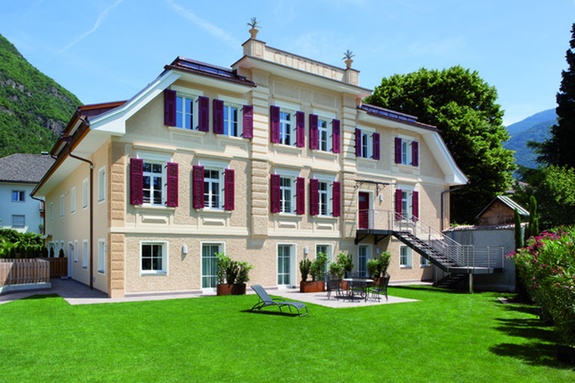 Elegante Villa mit hellgelber Fassade, roten Fensterläden, grünem Rasen und Bergen.