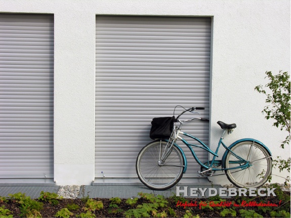 Türkisfarbenes Fahrrad lehnt an weißem Gebäude mit geschlossenen Rollläden.