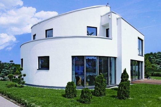 Weißes, modernes Haus mit gebogener Front, großen Fenstern und gepflegtem Garten unter blauem Himmel.
