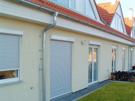 Moderne Hausfassade mit beigen Wänden, roten Ziegeln, Rollläden und grünem Rasen.