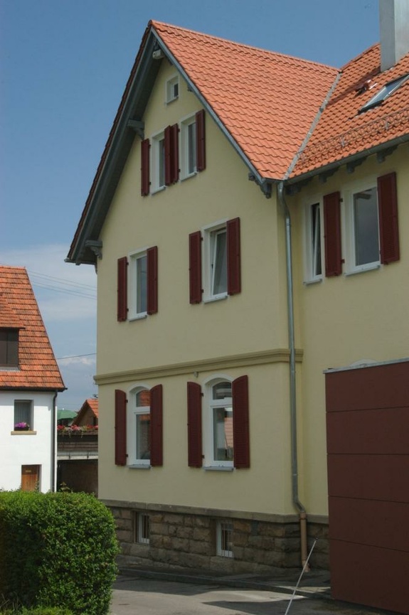 Helles gelbes Haus mit rotem Ziegeldach und Fensterläden unter blauem Himmel.