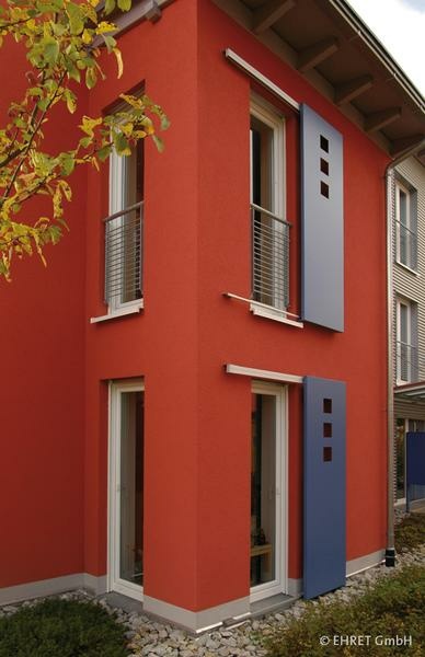 Modernes zweistöckiges rotes Haus mit blauen Schiebeläden, Balkon und Baum.