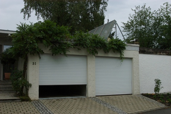 Doppelte Garage mit zwei weißen Rolltoren; das linke ist teilweise geöffnet. Efeu bewächst das Dach.