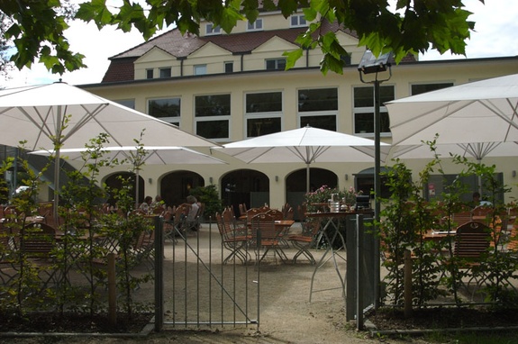 Sonnige Restaurantterrasse mit weißen Sonnenschirmen, besetzten Tischen und Gebäude.