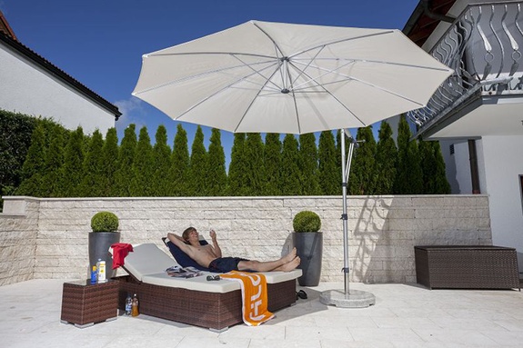 Mann entspannt sich auf Liege unter weißem Sonnenschirm auf sonniger Terrasse.