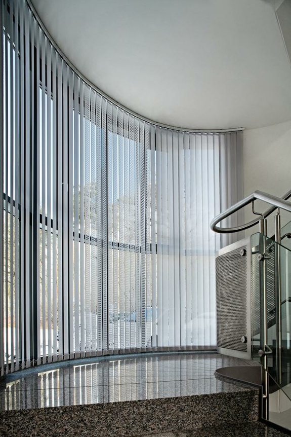 Moderne Innenansicht mit gebogener Fensterfront, vertikalen Jalousien und glänzendem Boden.