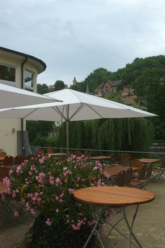 Gemütlicher Café-Außenbereich mit Holztischen, weißen Sonnenschirmen und rosa blühenden Pflanzen.