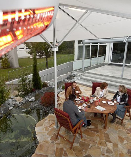 Vier Personen speisen auf einer Steinterrasse mit Teich, unter Schirm und rotem Heizstrahler.