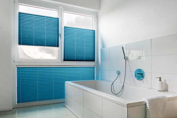 Helles Badezimmer mit weißer Badewanne und blauen Plissees am Fenster.