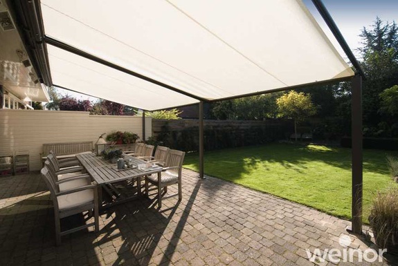 Sonnige Terrasse mit Esstisch und cremefarbener Pergolamarkise vor grünem Rasen.