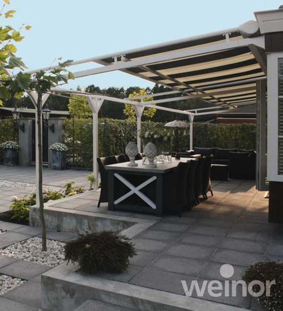 Moderne Terrasse mit weißer Pergola, Esstisch und Gartenmöbeln auf grauen Steinplatten.