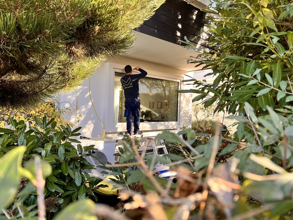 Mettler Techniker auf Leiter montiert ein Fenster an einem weißen Haus, umgeben von grünen Büschen.