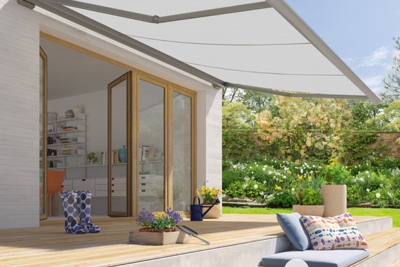 Fotografie: Helle, ausfahrbare Markise über einer Holzterrasse mit Blumen und grünem Garten.